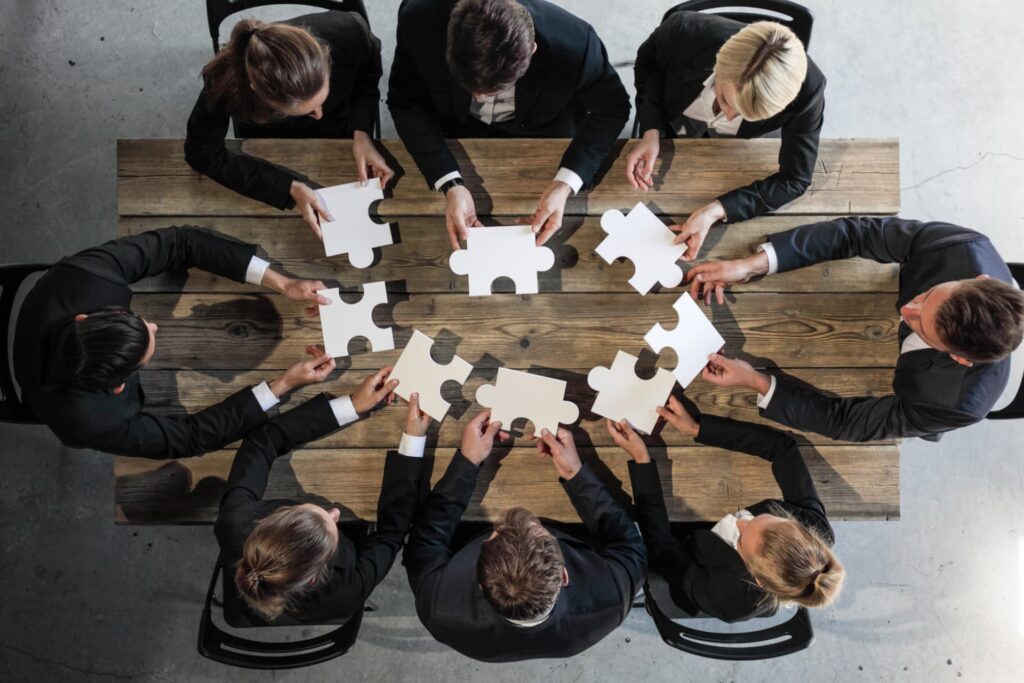 Group of professionals around a table assembling puzzle pieces, symbolizing collaboration among lawyers, accountants, mortgage brokers, realtors, and other insolvency specialists working with clients in financial stress.